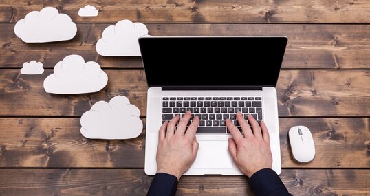 Cloud computing technology concept with white fluffy clouds next to the laptop. Mans hands typing the the keyboard uploading data, on a wooden table.